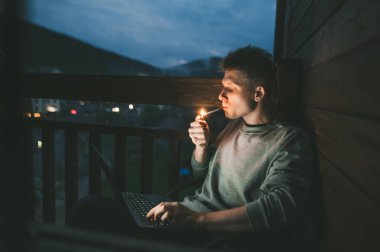 Handsome young man sits at night on the balcony with a laptop on his lap and lights a cigarette with a lighter.Evening portrait freelancer smokes and works on a laptop on a balcony in an apartment clipart