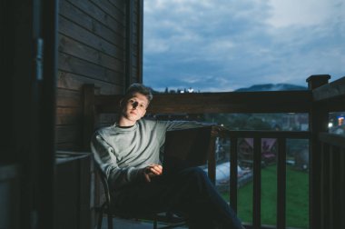 Evdeki balkonda dizüstü bilgisayarda çalışan yakışıklı, serbest çalışan genç bir adamın akşam portresi. Ciddi bir yüzle kameraya bakıyor. Bilgisayarlı adam otelin balkonunda oturuyor..