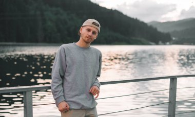 Gri kazaklı ve şapkalı bir adam gölün ve köknar ormanının manzarasının tadını çıkarıyor. Genç bir turist dağ ormanlarındaki göletin yakınındaki kameraya bakıyor. Akşam manzarası.