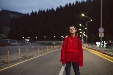 Genç bir gezgin kızın portresi. Akşamleyin dağ ormanının yanında yolda yürüyor. Güzel bayan turist, dağlık ormanın yakınlarında geziniyor. Sokakta kendini rahat ve huzurlu hissediyor..