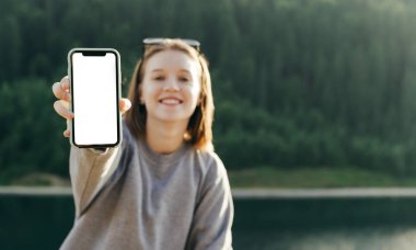 Gülümseyen kadın yolcunun dağ ormanlarının arka planında akıllı telefon boş ekranı gösteren portresi. Gülümseyen neşeli turist kız köknar ormanı manzarasında telefon ekranını gösteriyor.