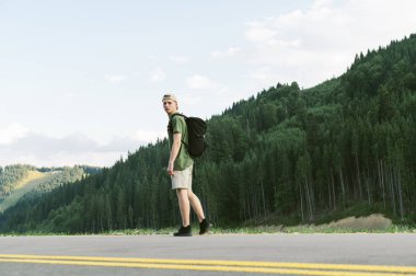 Genç erkek turist dağ manzarasında yolda duruyor. Çekici gezgin adam dağ kozalaklı ormanların yakınındaki kameraya bakıyor. Peyzaj kompozisyonu. Yaşam tarzı, seyahat kavramı.