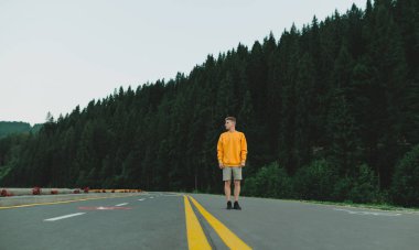 Dağlardaki köknar ormanlarının arka planında yolda duran düşünceli beyaz erkek yürüyüşçü. Ciddi sakin genç gezgin, kenara bakıyor, ormanlık alanda otoyolda poz veriyor..