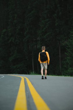 Sarı kazaklı erkek turist, köknar ormanlarının arka planında asfalt yolda yürüyor. Dikey çekim, dağ ormanının yakınındaki otobanda gezgin adamın arka görüntüsü..