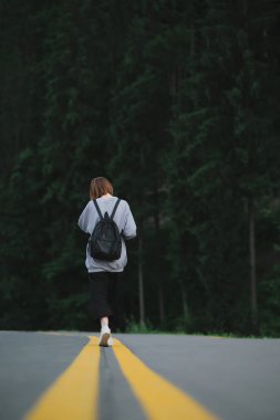 Gri kazaklı, siyah pantolonlu, sırt çantalı, dağ ormanlarında asfalt yolda gezinen kadın gezginin dikey görüntüsü. Turist kızın, kozalaklı ormanın yakınlarında yürüdüğünü gördüm..