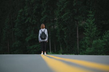 Dişi gezginin gece vakti orman arka planında yürürken çekilmiş yatay görüntüsü. Ormanın yakınındaki asfalt yolda sırt çantalı bir turist kız..