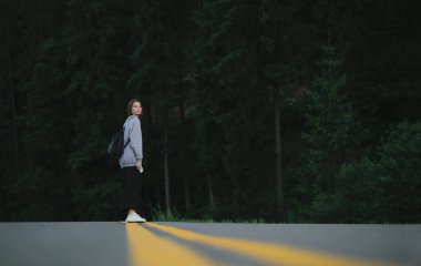 Genç bayan gezgin, dağ ormanlarında asfalt yolun ortasında kameraya bakarak duruyor. Neşeli turist kadın, köknar ormanları arasında yolun ortasında kameraya poz veriyor..