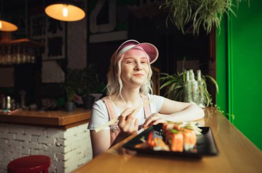 Parlak bir makyaj ve pembe giysiler ile Mutlu bayan bir restoranda oturur ve yemek çubukları ile suşi rulo yiyor, kamera içine bakar ve gülümsüyor. Mutlu kız Japon yemekleri yemek ten hoşlanıyor.