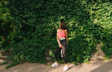 Arka planda, sarmaşık duvarıyla dans eden mutlu İspanyol kız. Tarz sahibi kıvırcık saçlı melez kız yeşil bir duvarın arka planında eğleniyor, gülümsüyor ve başka tarafa bakıyor..