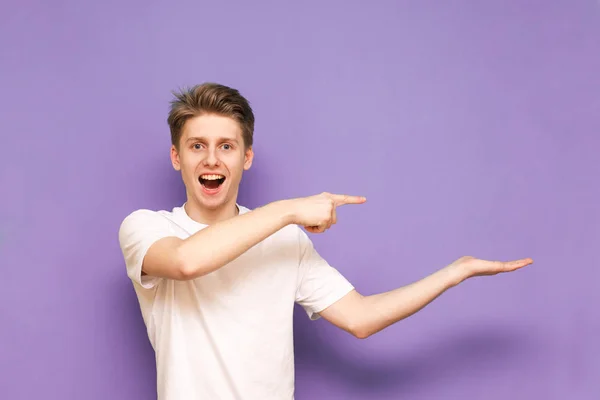 Jovem feliz em uma camiseta branca fica em um fundo roxo e mostra com as mãos e uma missão vazia para publicidade, sorri e olha para a câmera. Isolado. Espaço de cópia — Fotografia de Stock