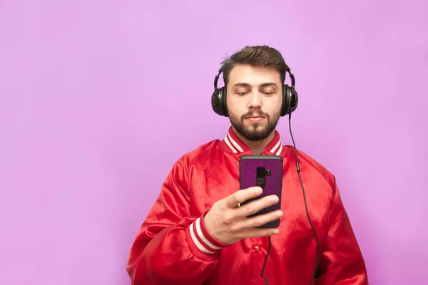 Retrato de un hombre barbudo en los auriculares de pie sobre el fondo de una pared rosa con un teléfono inteligente en la mano, mirando enfocado en la pantalla, usando una chaqueta roja. Aislado . —  Fotos de Stock