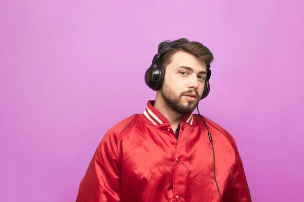 Retrato de um homem com barba, vestindo uma jaqueta vermelha, fica no fundo de uma parede rosa, e ele ouve música nos fones de ouvido. . — Fotografia de Stock