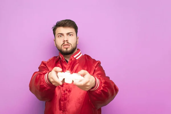 Retrato de um jogador concentrado com uma barba em uma jaqueta vermelha fica em um fundo rosa com um joystick na mão e sofisticado olhando para a câmera, jogando videogames. Conceito de jogador — Fotografia de Stock