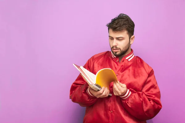 Retrato de un estudiante americano con una barba de pie sobre un fondo rosa con un cuaderno en la mano y lecturas. Hombre con libros en la mano y una chaqueta roja, aislado sobre un fondo de una pared rosa — Foto de Stock