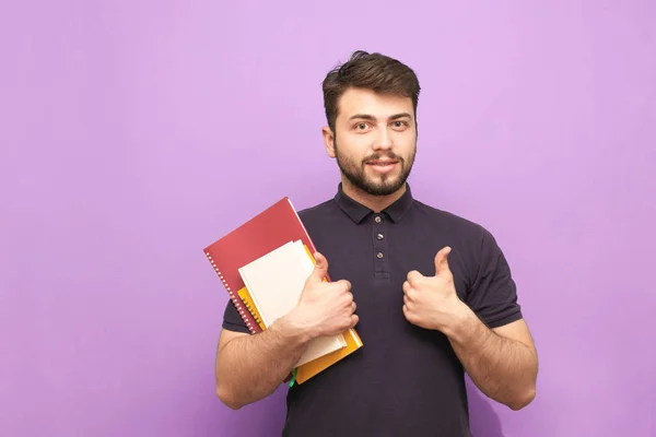 Портрет бородатого студента-мужчины, стоящего с книгами и тетрадками в руках, улыбающегося и указывающего пальцем в камеру. Счастливый парень с книгами позирует перед камерой . — стоковое фото