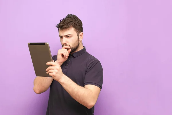 Un hombre concentrado con una barba y una camiseta se levanta sobre un fondo púrpura y mira la pantalla con cuidado, con una camiseta oscura. Aislado. Hombre de negocios utiliza una tableta . — Foto de Stock