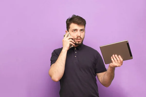 Portrait of a busy man standing on a blue background with a tablet in his hands and talking on the phone.Business man with gadgets in his hands is isolated on a purple background. Copyspace