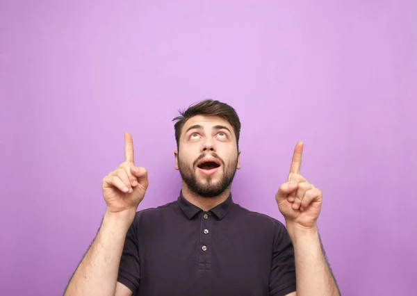 Portrait of a surprised man on a purple background, looking up and showing his fingers on a blank space. Man with a beard shows hands on copyspace, isolated