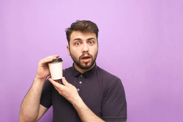 Удивленный мужчина в тёмной рубашке и с бородой, держа в руках бумажную чашку кофе и глядя на камеру на фиолетовом фоне с эмоциональным лицом. Isolated . — стоковое фото