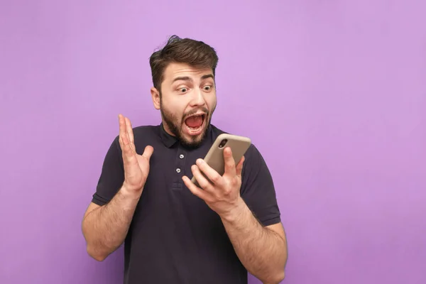 Portrait of a crazy bearded man with a smartphone in his hands, looking at the screen and screaming on a purple background. Shocked man uses a smartphone and rejoices. Isolated. — Stock Photo, Image