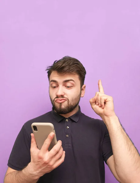 Emotional man with a beard holds a smartphone in his hands, showing his finger up in a blank space. Surprised adult in a dark T-shirt uses a smartphone and shows a finger on copyspace. purple backgrou — Stock Photo, Image