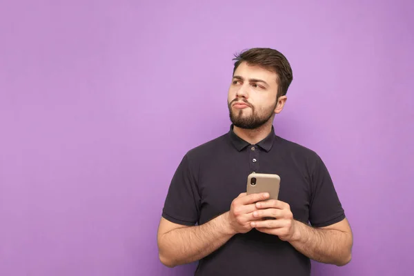 Retrato de un hombre desconcertado en la ropa casual se encuentra con un teléfono inteligente en sus manos, mirando pensativamente hacia los lados sobre un fondo púrpura. Espacio de copia — Foto de Stock