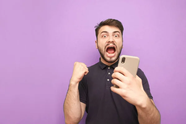 Emotional bearded man holds the smartphone in his hands, looks at the camera and rejoices. Man with a smartphone in his hands rejoices in success, isolated on a purple background. Copyspace — Stock Photo, Image