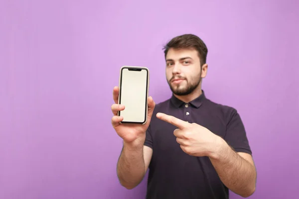Smiling man holds a smartphone in his hands, and shows his finger on a white screen on a pink background. Bearded man wears a dark T-shirt and shows a smartphone with a white screen in the camera. — Stock Photo, Image