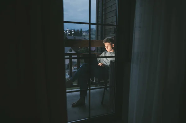 Συγκεντρωμένος ελεύθερος επαγγελματίας κάθεται το βράδυ στο μπαλκόνι του διαμερίσματος εργάζονται σε ένα φορητό υπολογιστή, κοιτάζοντας έντονα στην οθόνη. Νεαρός χρησιμοποιεί φορητό υπολογιστή τη νύχτα στο μπαλκόνι στο σπίτι. Έννοια της ελευθερίας. — Φωτογραφία Αρχείου