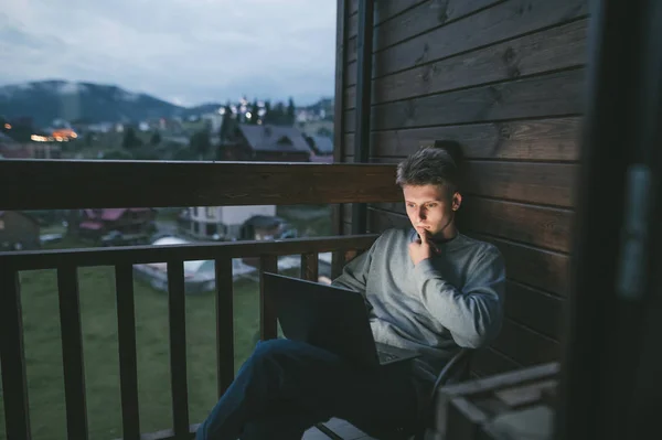 Guy κάθεται στο εξοχικό σπίτι στο μπαλκόνι το βράδυ και χρησιμοποιώντας ένα φορητό υπολογιστή, κοιτάζοντας προσεκτικά την οθόνη.Νεαρός ελεύθερος επαγγελματίας που εργάζονται σε φορητό υπολογιστή σε εξοχικό σπίτι στο μπαλκόνι με όμορφη θέα το βράδυ — Φωτογραφία Αρχείου