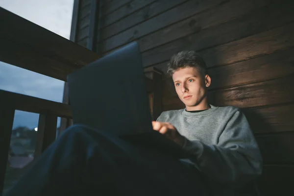 Χαριτωμένος τύπος που κάθεται το βράδυ σε ένα ξύλινο μπαλκόνι ενός εξοχικού σπιτιού με ένα φορητό υπολογιστή στην αγκαλιά του και εργάζεται, κοιτάζοντας την οθόνη του υπολογιστή. Ελεύθερος επαγγελματίας που εργάζεται το βράδυ σε ένα φορητό υπολογιστή — Φωτογραφία Αρχείου