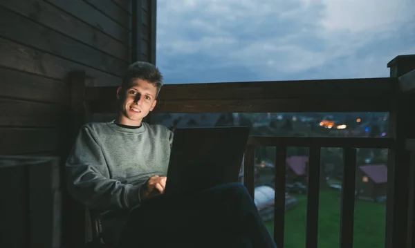 Happy young man sitting on balcony in country house in the evening with laptop in hands, looking into camera and smiling.Smiling freelancer working late at night on laptop in country apartment