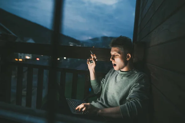 Young man smokes a cigarette on the balcony and uses a laptop in the evening at a country house,looks into the screen. Guy sits on the balcony in a chair with a cigarette and works on a laptop
