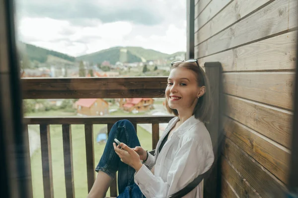 Happy girl in stylish casual clothes and with smartphone in her hands sits on the balcony in the cottage, looks into the camera and smiles. Smiling woman relaxing on balcony in home outside city.