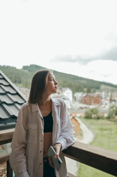 Retrato de menina atraente em pé na varanda no hotel em montanhas, olhando para longe com rosto sério. Mulher bonita em roupas elegantes relaxando em um campo nas montanhas. Foto vertical . — Fotografia de Stock