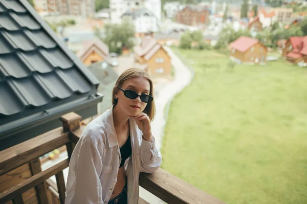 Menina elegante posando na varanda no quarto do hotel, usa óculos de sol, olha para a câmera com um rosto sério. Mulher bonita de pé na varanda na casa de campo em montanhas . — Fotografia de Stock