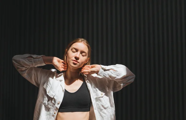 Portret van mooi meisje in wit shirt en donkere beha op donkere achtergrond, poseren met gesloten ogen, licht vangen en genieten. Kopieerruimte — Stockfoto