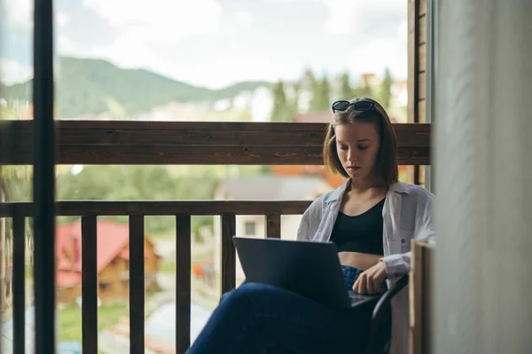 Koncentrált, stílusos nő ül az erkélyen egy laptoplakásban és dolgozik, mereven bámulja a képernyőt. Gyönyörű lány szabadúszó dolgozik egy laptopon egy vidéki házban. Szabadúszó — Stock Fotó