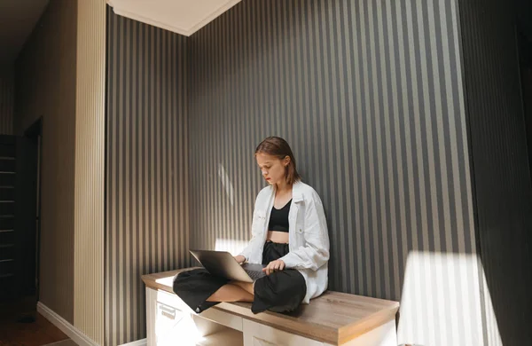 Menina atraente sentado no apartamento com o laptop em seu colo e trabalho, foco olha para a tela do computador. Retrato de mulher freelancer trabalhando em casa com laptop . — Fotografia de Stock