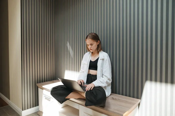 Стильна приваблива дівчина сидить на трибуні стильна квартира і використовує ноутбук. Портрет фрілансера, який працює вдома та в Інтернеті. Серйозна дівчина-фрілансер використовує Інтернет — стокове фото