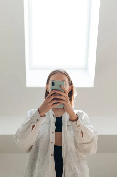 Mulher em roupas casuais elegantes, vestindo uma camisa branca fica com um smartphone em suas mãos em apartamentos em uma janela de sótão leve, cobre seu rosto com um smartphone. Retrato dentro de casa . — Fotografia de Stock
