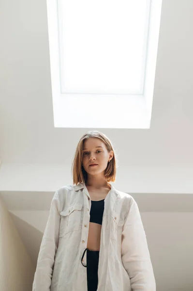 Chica sexy en una camisa superior y blanca posa en un ático ligero apartamento, mira a la cámara. Contra la ventana del ático en el techo. Foto vertical . — Foto de Stock