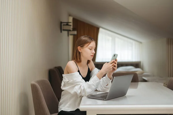 Elegante chica sentada en un escritorio con un ordenador portátil en apartamentos con un elegante interior de luz, utilizando un teléfono inteligente. Mujer joven freelancer trabajando en casa en el portátil y el teléfono con ropa casual con estilo — Foto de Stock