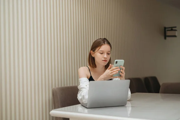 Hermosa chica de negocios utiliza teléfono inteligente y portátil en la mesa en casa en la mesa en el apartamento. Mujer atractiva en ropa casual trabajando en casa con un ordenador portátil, retrato en la habitación. Independiente — Foto de Stock