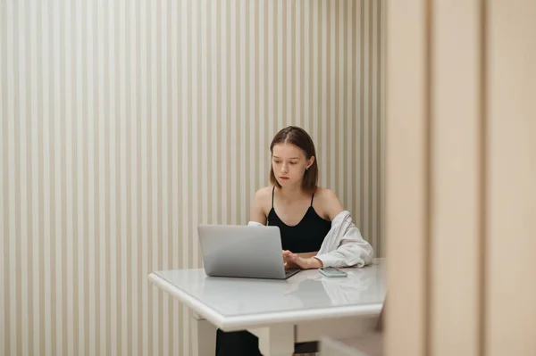 Chica atractiva se sienta en una mesa en casa y utiliza un ordenador portátil, mira atentamente a la pantalla, lleva ropa casual con estilo. Estudiante estudia en su habitación en la mesa, utiliza un ordenador portátil . — Foto de Stock