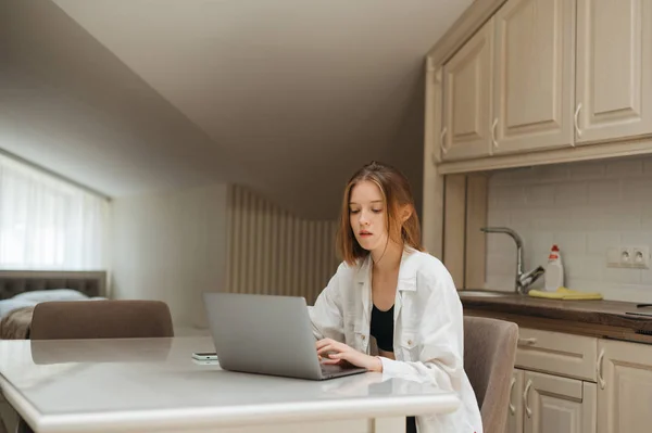 Красива дівчина сидить на кухні в квартирі, працює на ноутбуці, виглядає зосередженою на екрані. — стокове фото