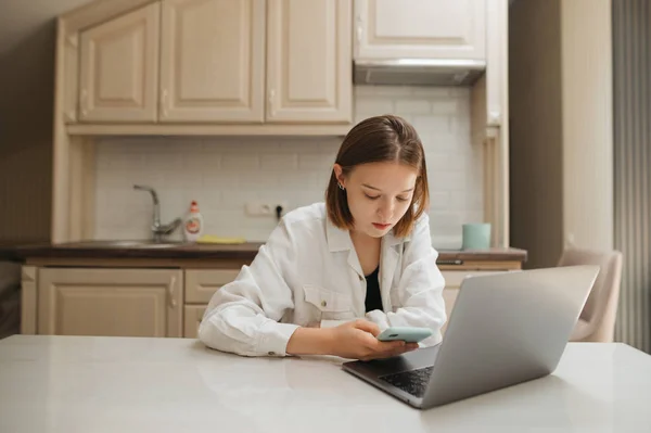 Красива дівчина в білій сорочці сидить з ноутбуком за столом на кухні і використовує смартфон. Симпатична дівчина працює на гаджетах вдома в квартирі . — стокове фото