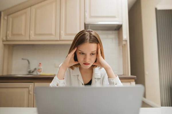 Λυπημένη μαθήτρια που σπουδάζει στο σπίτι. Δυστυχισμένη κοπέλα κάθεται στο τραπέζι στην κουζίνα σε διαμέρισμα με φορητό υπολογιστή και κοιτάζοντας την οθόνη με ζοφερή πρόσωπο.Θυμωμένη κοπέλα λειτουργεί σε ένα φορητό υπολογιστή στην κουζίνα — Φωτογραφία Αρχείου