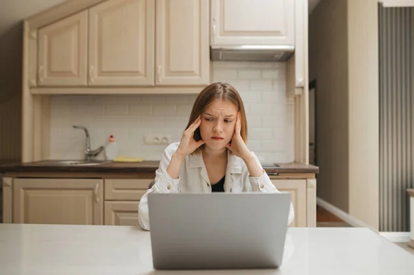 Ντροπιασμένη ελκυστική κοπέλα που δουλεύει σε ένα λάπτοπ στο τραπέζι της κουζίνας, κοιτάζοντας αλλού με θλιμμένο πρόσωπο. Πορτρέτο ενός χαρούμενου κοριτσιού χρησιμοποιώντας ένα φορητό υπολογιστή στην κουζίνα στο διαμέρισμα. — Φωτογραφία Αρχείου