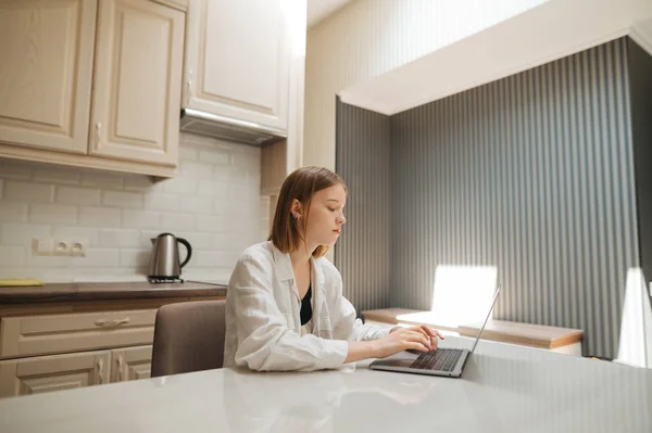 キッチンのテーブルに座ってノートパソコンを使っているフリーランスの女の子の肖像画で、画面と作業を真剣に見ています。白いシャツの美しい女性はアパートでラップトップを使用しています — ストック写真